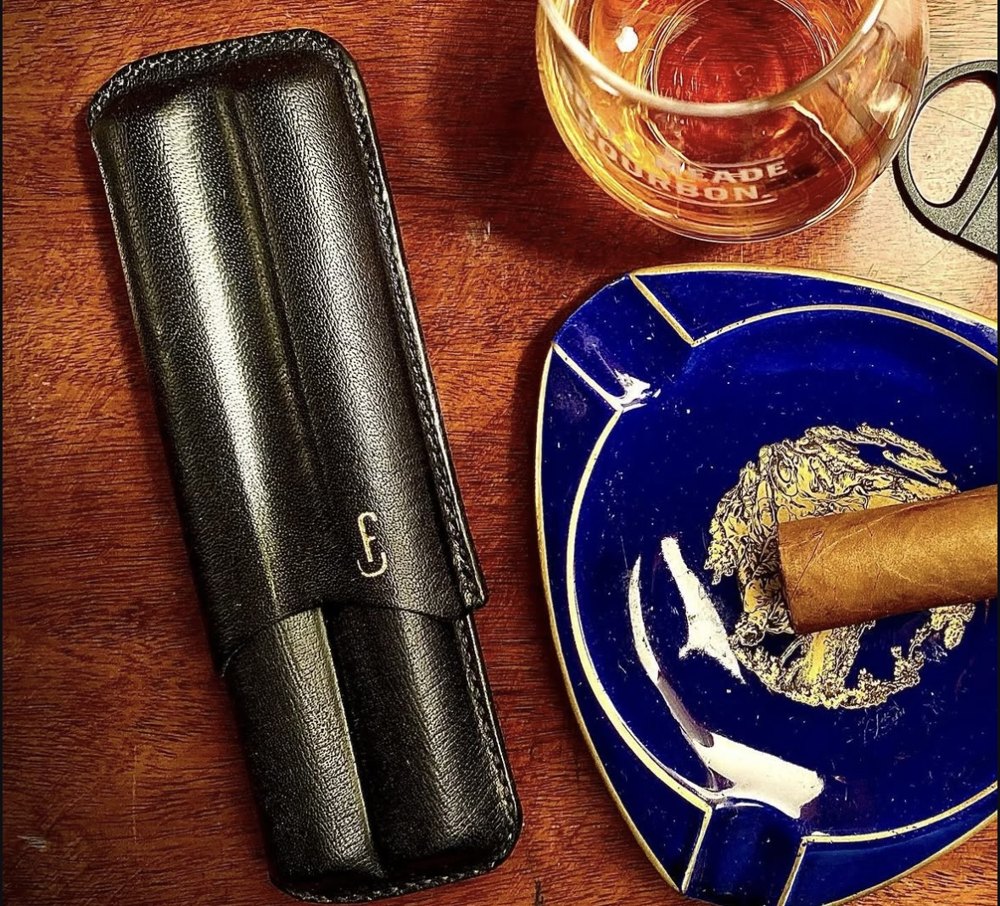 A leather cigar case beside whiskey and a cigar